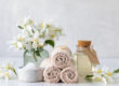 Spa essentials with rolled beige towels, bath salt in white speckled bowl, jasmine flowers in glass vase, and essential oil in glass bottle on a light background.