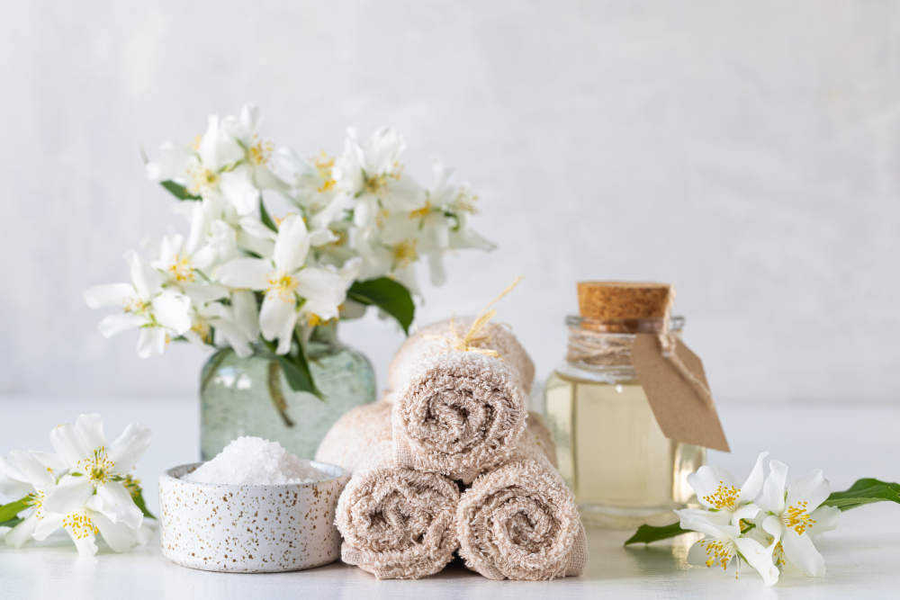 Spa essentials with rolled beige towels, bath salt in white speckled bowl, jasmine flowers in glass vase, and essential oil in glass bottle on a light background.