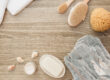 Spa and wellness setup featuring a white towel, natural bristle body brush, sea sponge, bar of soap, open jar of cream, and seashells on a wooden textured background.