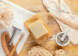 Zero waste bathroom accessories on a yellow textured background, including a white towel, wooden soap dish with bar soap, bamboo toothbrushes, body brush, loofah, safety razor, and a mortar with grey powder.