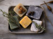 Assorted artisanal soaps on a rustic tray, featuring ingredients like pecan, charcoal, and herbs, alongside a wooden spinning top and fresh rosemary sprigs, set on a textured grey surface.