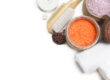 Spa essentials with bath salts in wooden bowls, a white candle, towel, and natural brushes on a white background.