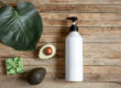Natural skincare setup with a large white lotion dispenser, half an avocado, a green handcrafted soap, and a big green leaf on a rustic wooden background.